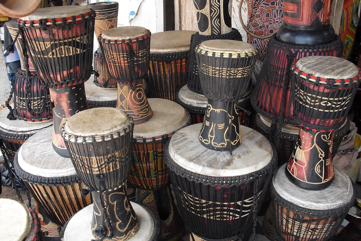 A crowded display of djembe drums with carved wooden shells, rope tuning and pale drumheads, on sale at a craft village